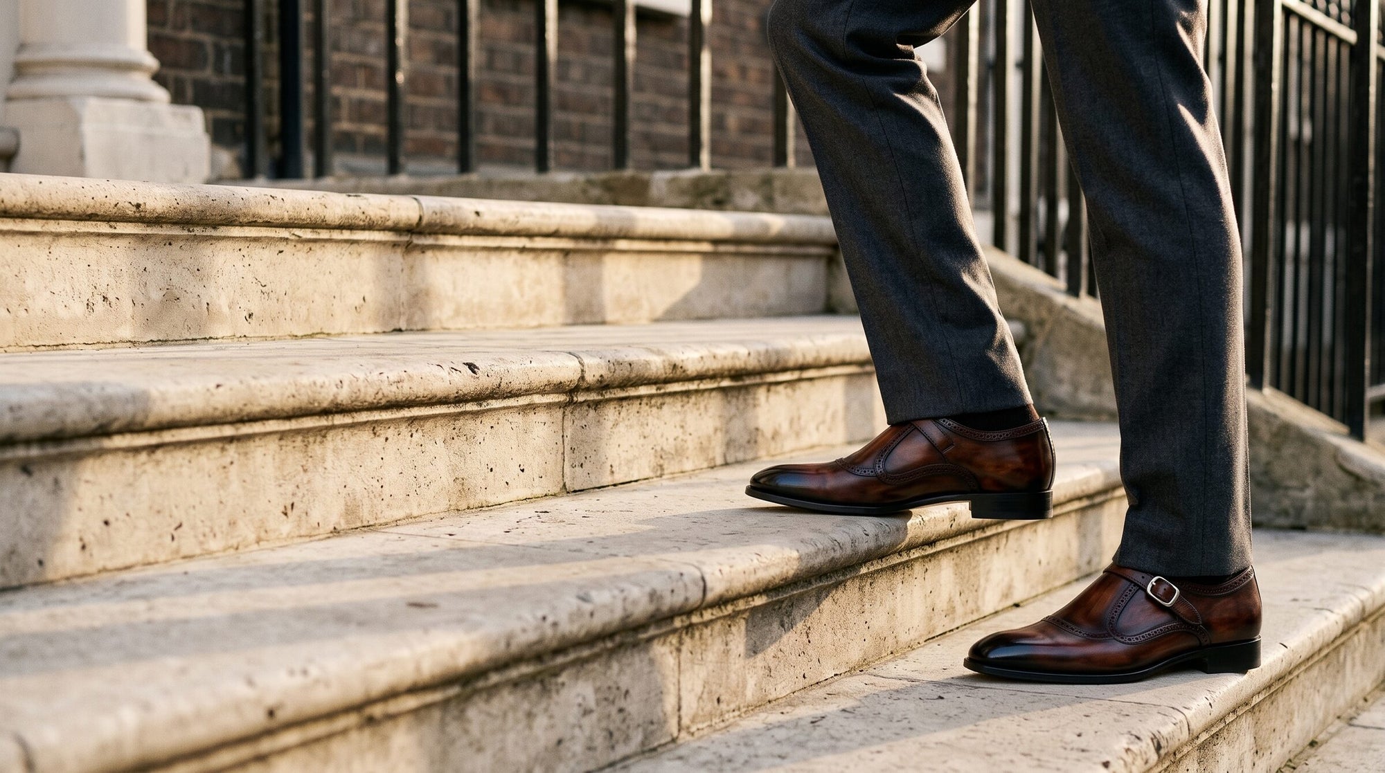 Brown patina monk strap shoes worn with charcoal trousers, climbing the stone steps of a London townhouse